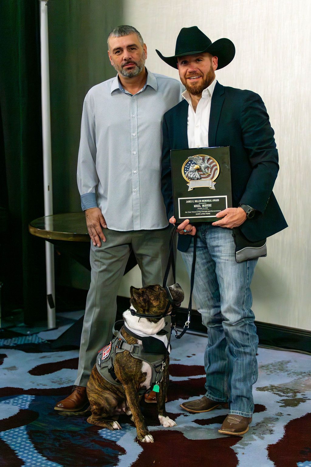 Two men stand with a brindle dog wearing a harness; one man holds an award plaque in an indoor, carpeted room.