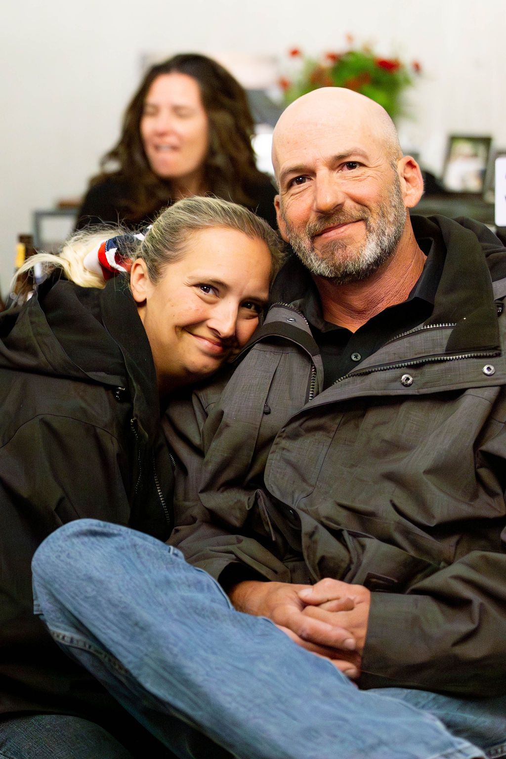 A smiling couple poses for a photo, with the woman resting her head on the man's shoulder. A blurry figure sits behind.