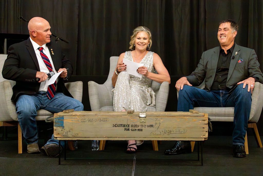 Three people sit in chairs on a stage with a wooden crate in front of them, engaged in a discussion.