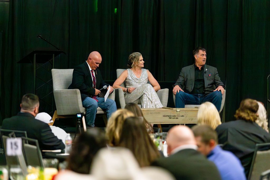 Three people sit on stage in armchairs during a panel discussion, facing an audience in a dimly lit conference room.