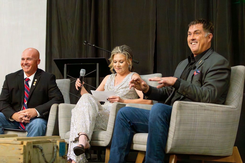 Three people sit in chairs on stage, one speaking with a microphone while others listen in an indoor event setting.