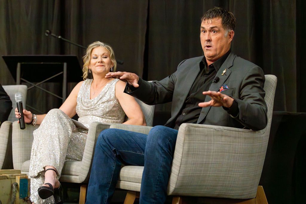 Two people in business-casual attire sit in chairs on a stage; one gestures while speaking, the other holds a microphone.