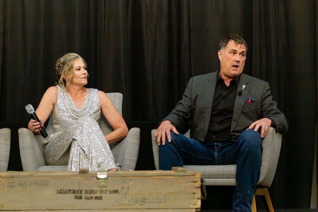 A person in a silver sequined outfit and a person in a gray blazer sit in armchairs on a stage, speaking into microphones.