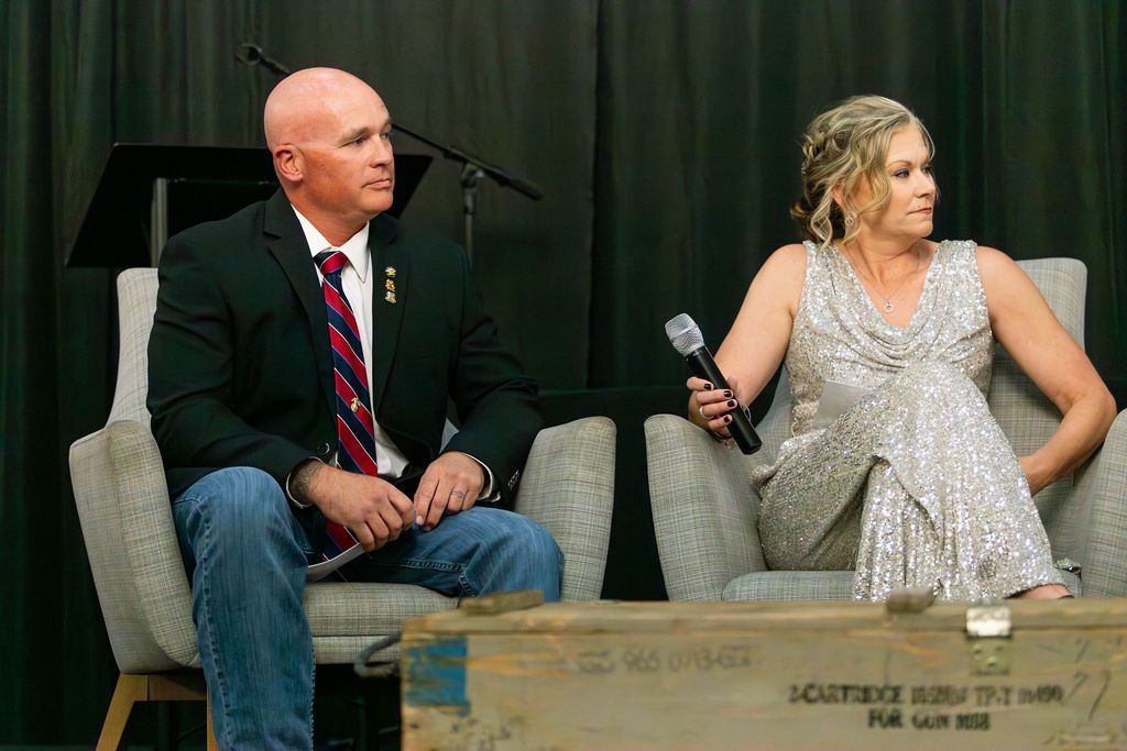A man in a suit and a woman in a formal gown sit in chairs on a stage with a microphone in front of a dark backdrop.