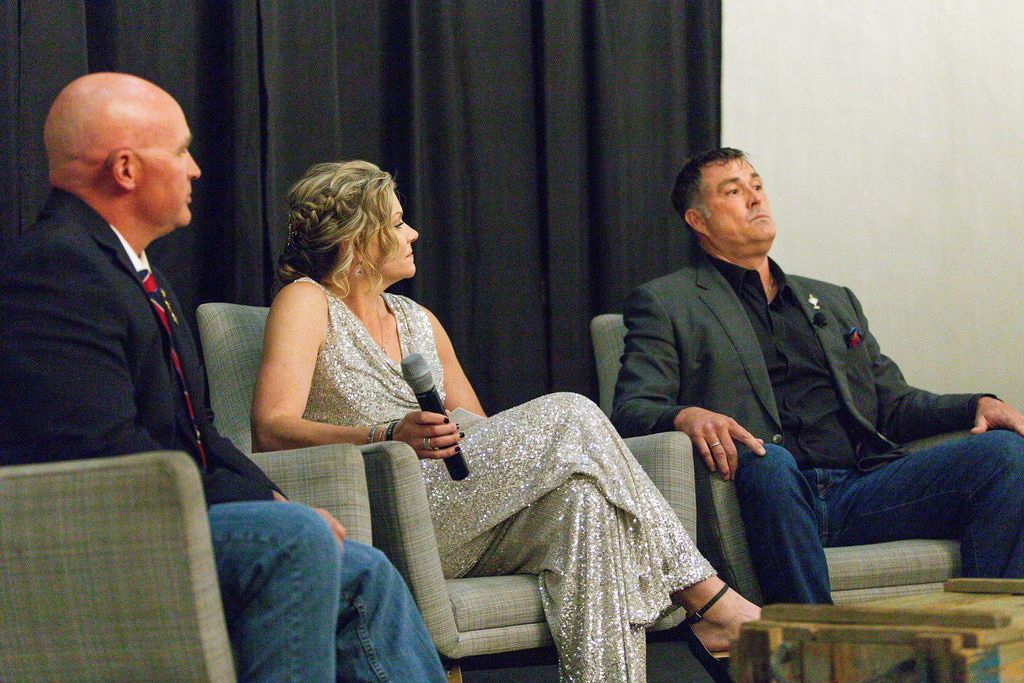Three people sit in chairs on a stage in front of a black curtain, with one person holding a microphone.