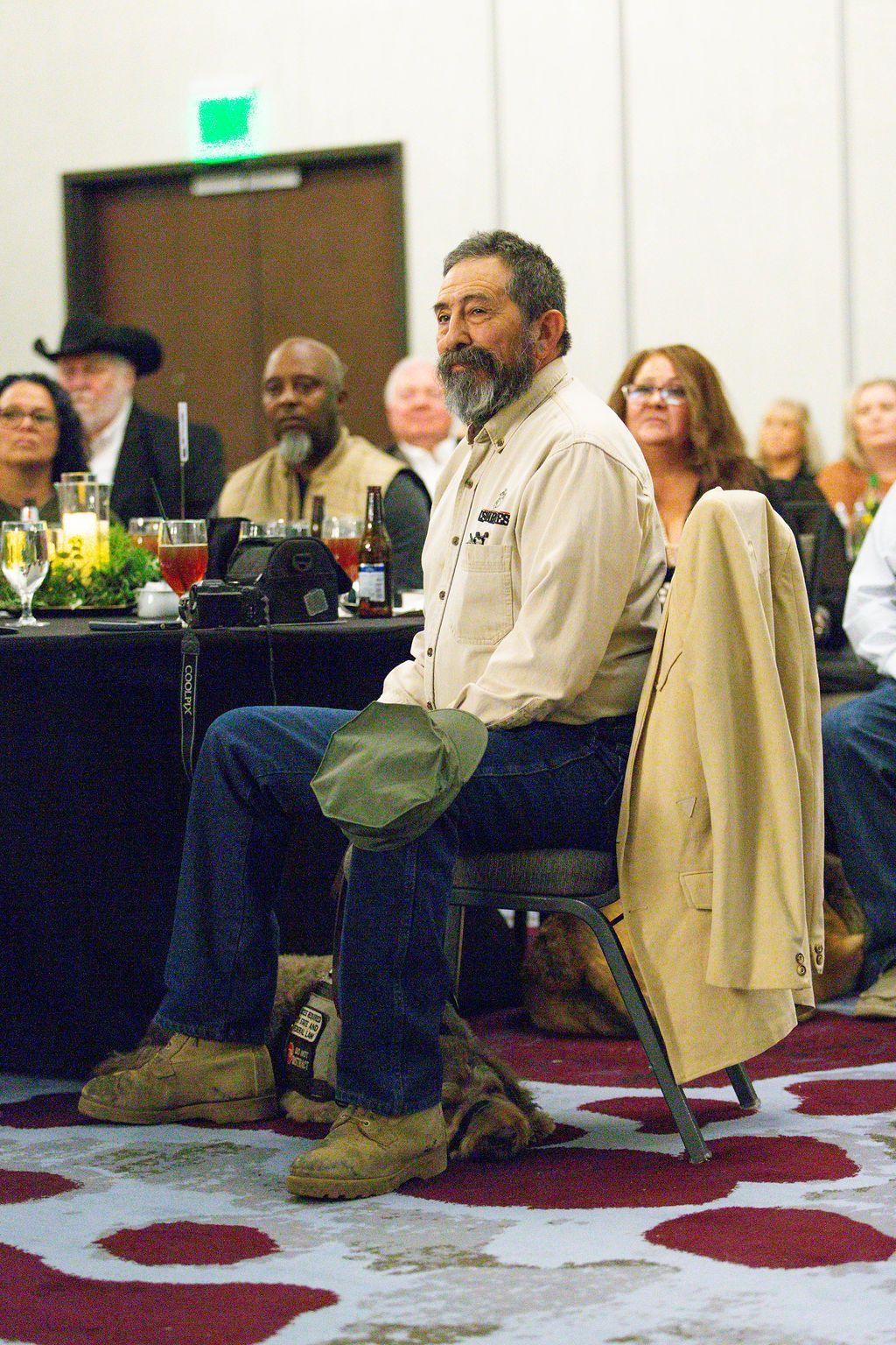 A person with a beard sits at a table in a conference room, wearing a light shirt, blue jeans, and tan boots.