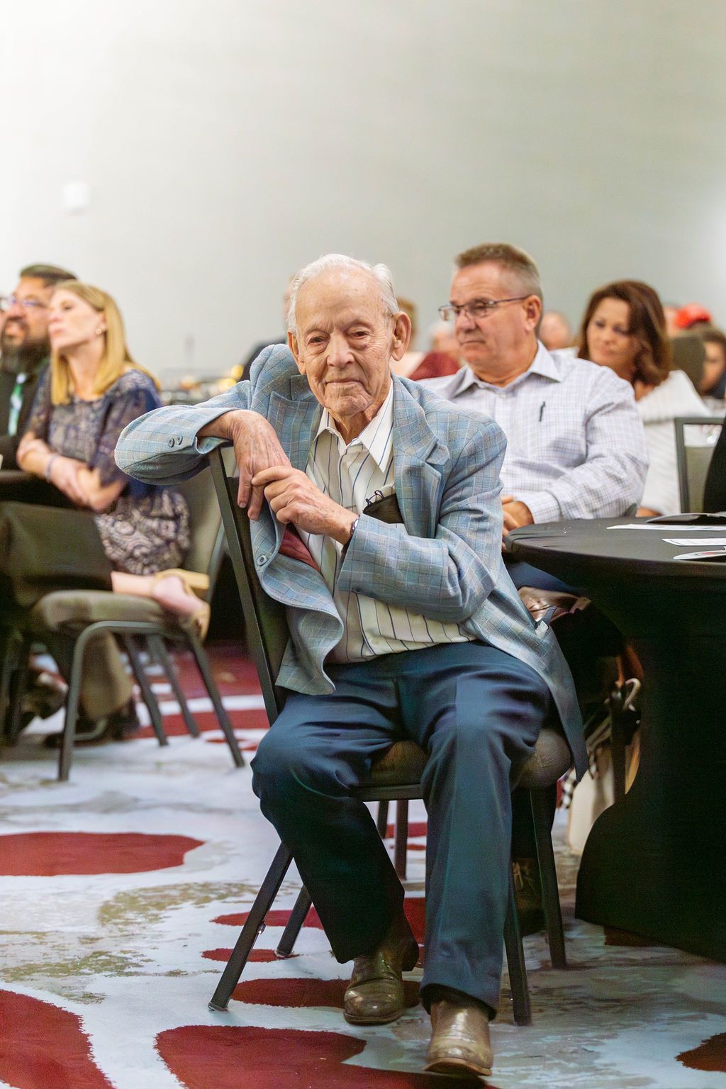 A man sits in a chair at a table in a conference room, with other attendees blurred in the background.