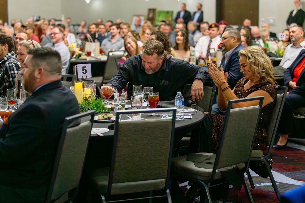 A group of people seated at round tables in a conference hall, with a man and woman appearing to toast or talk at a table.