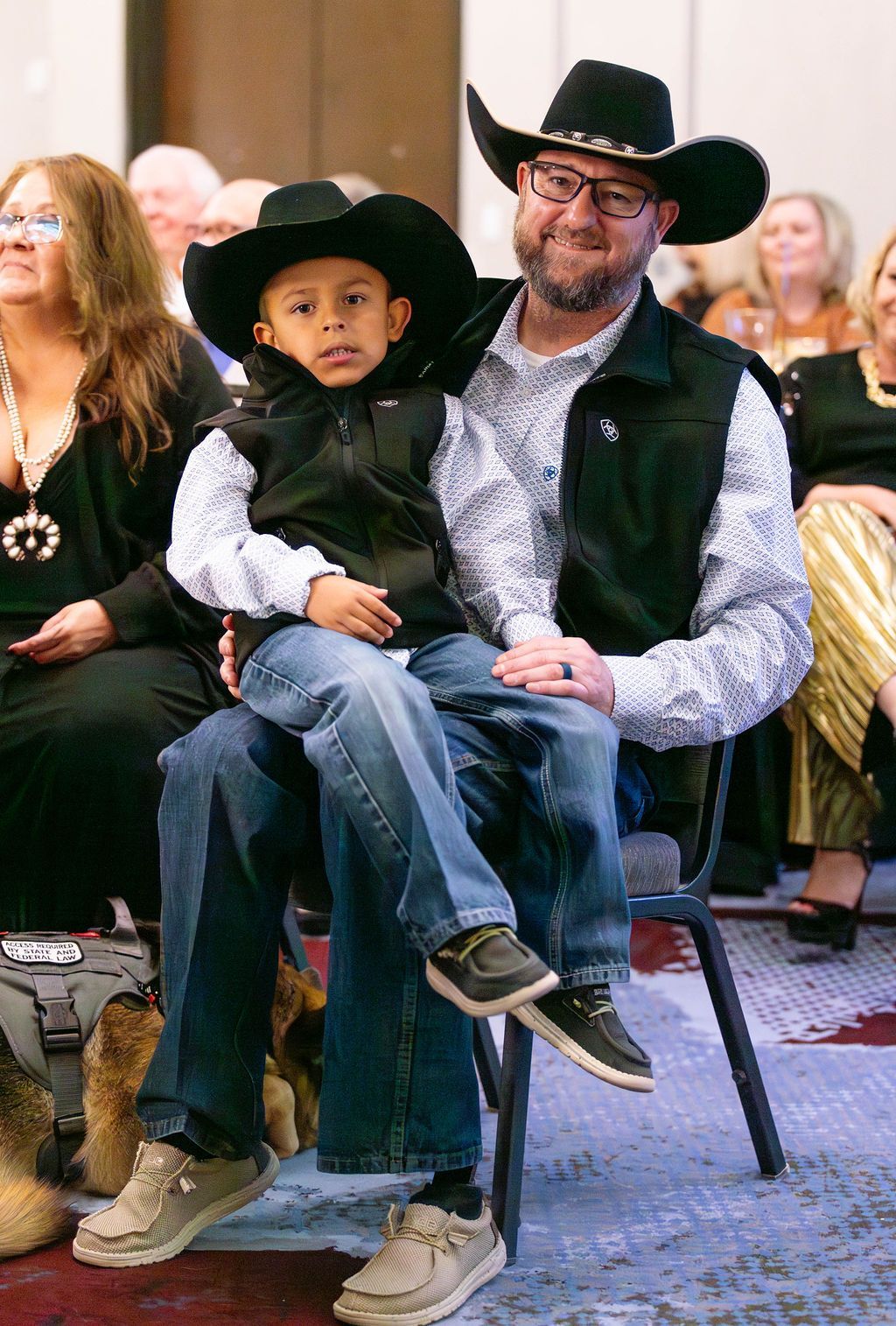 A person wearing a black cowboy hat and vest sits in a chair, holding a child on their lap who also wears a cowboy hat.