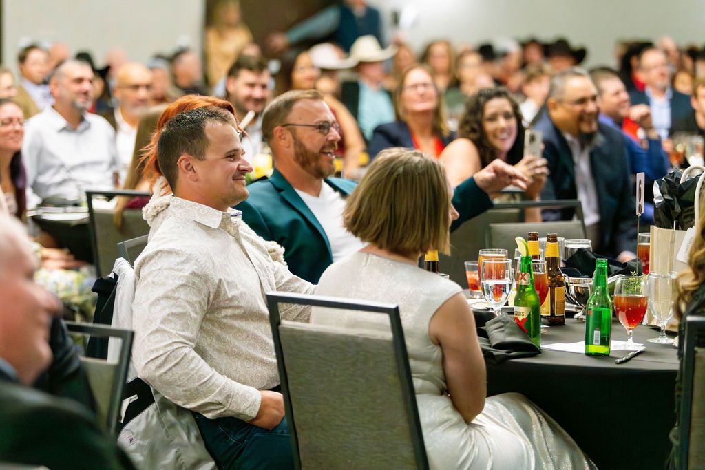 A group of people attending an event at dinner tables, with two individuals in the foreground smiling and conversing.