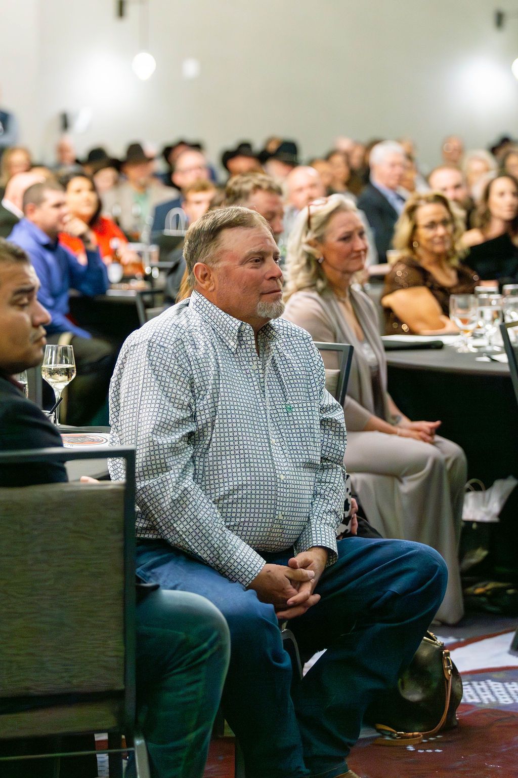 A group of people sit at round tables in a brightly lit banquet hall, focused attentively on a speaker off-camera.