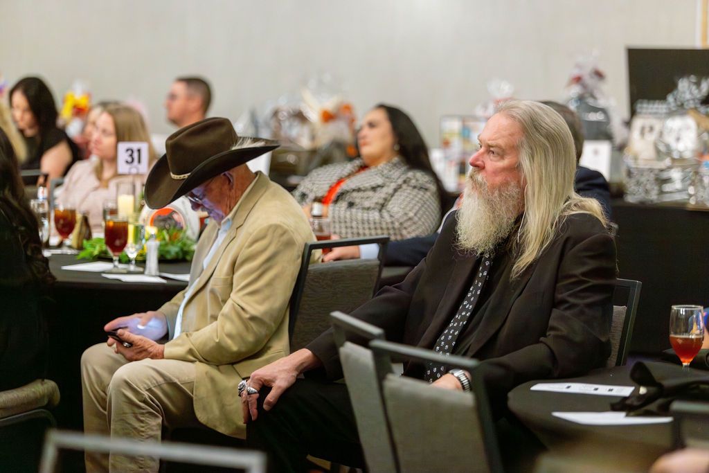 People seated at tables in a conference room, including a person with a beard and a person wearing a cowboy hat.