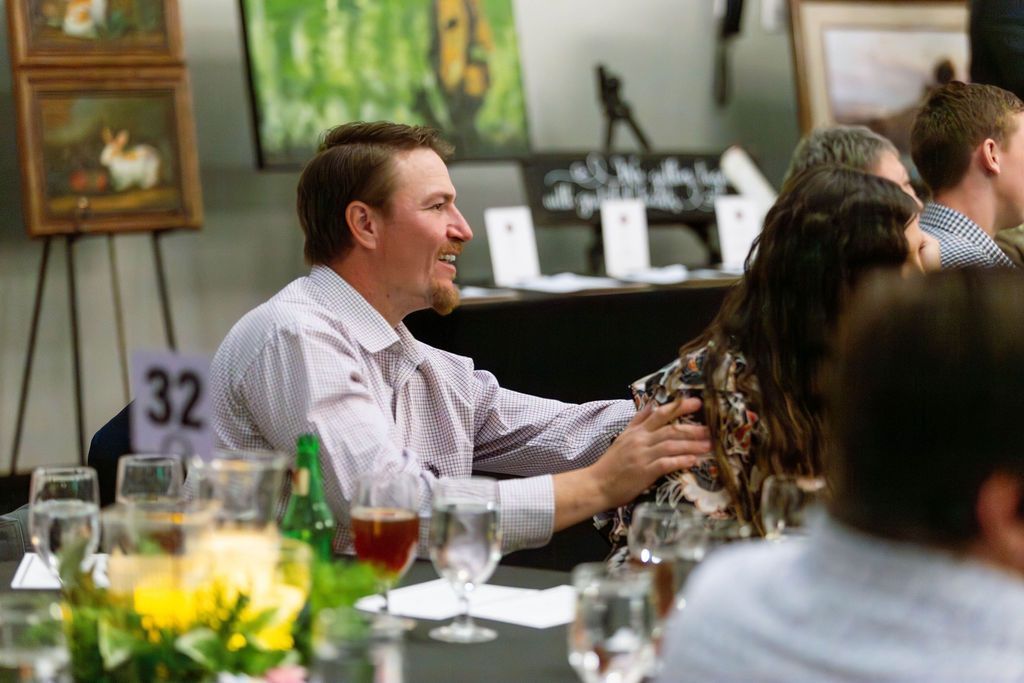 A man in a light-colored button-down shirt rests his hand on a person's shoulder at a table in a room with art on display.