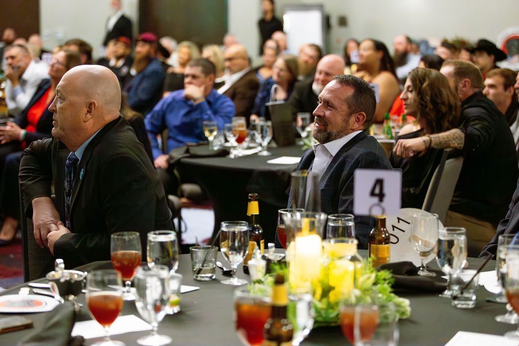 Attendees sit at round tables in a banquet hall, listening to a presentation during a professional event.