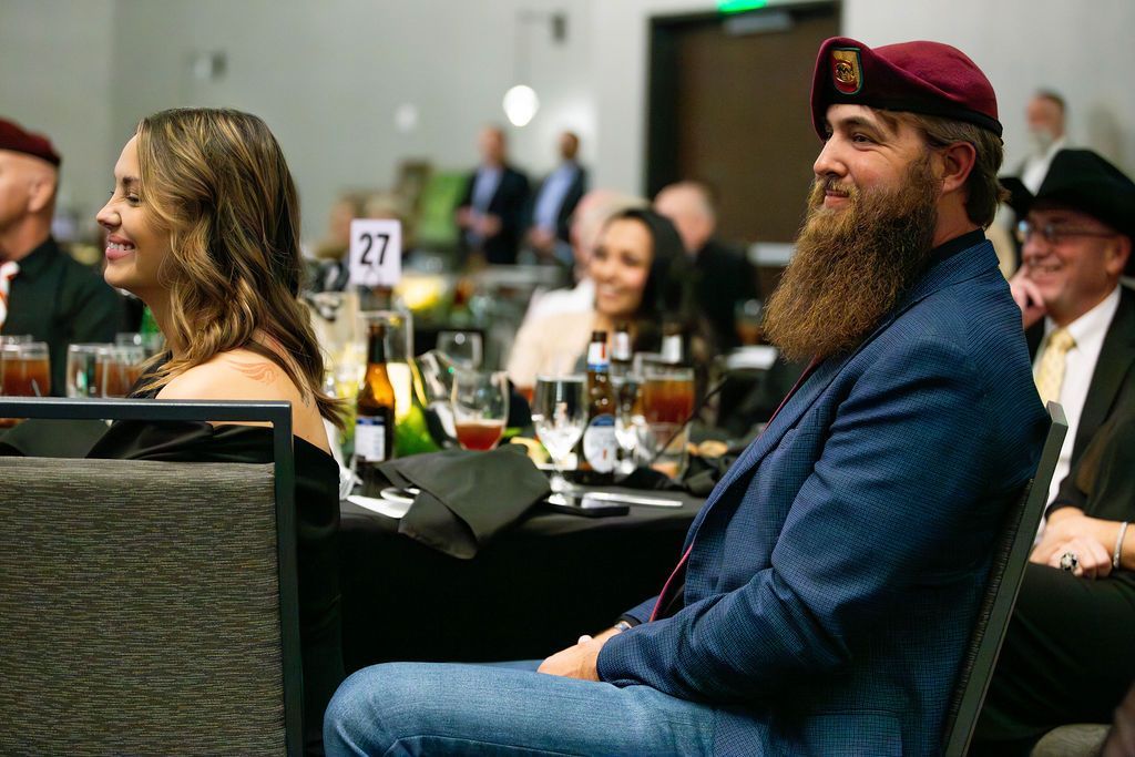 A group of people sit at tables in a banquet hall, with a bearded man in a maroon beret in the foreground.