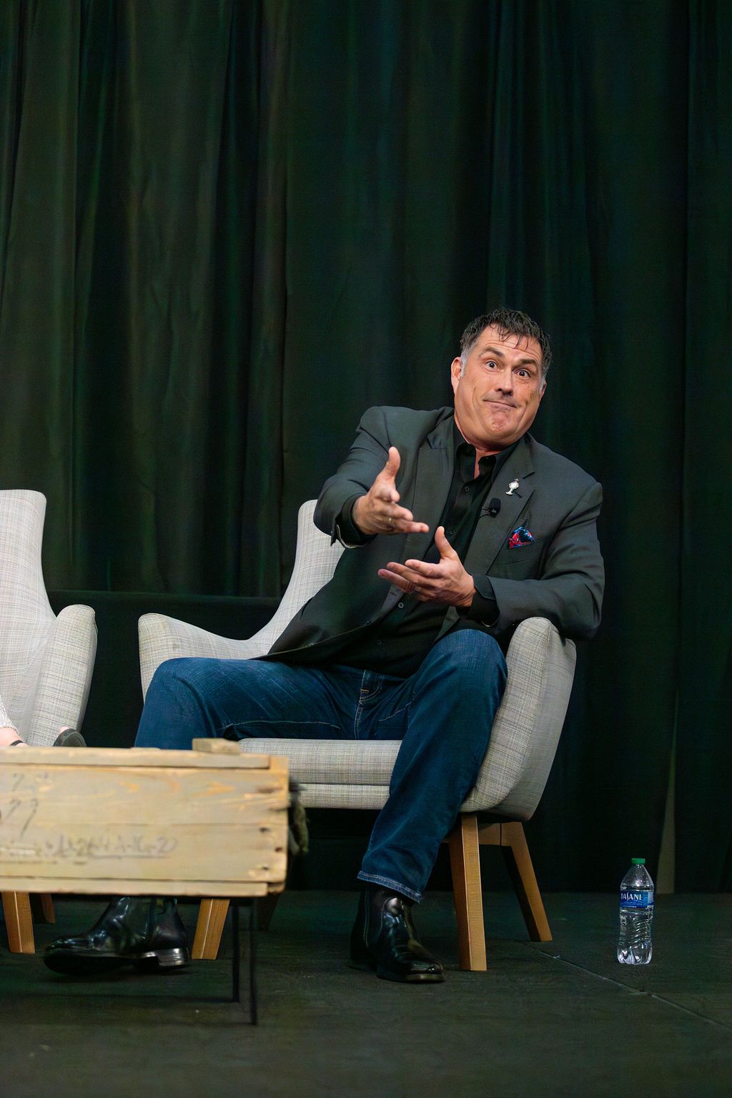 A person in a blazer and jeans sits in a chair on a stage, gesturing while speaking in front of a dark green curtain.