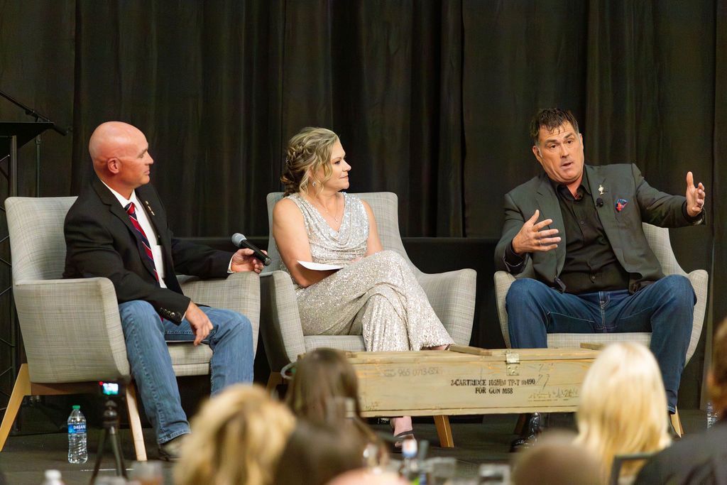 Three people sit in armchairs on a stage for a panel discussion in front of an audience, with a wooden crate in front.