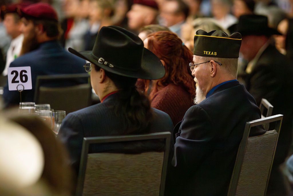 Attendees in hats and formal attire sit at a table marked with a number 26 at an indoor event.