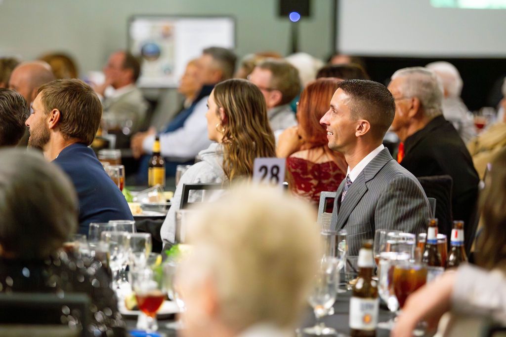 A side profile of people at a banquet, focusing on a person in a gray suit looking toward the front of the room.
