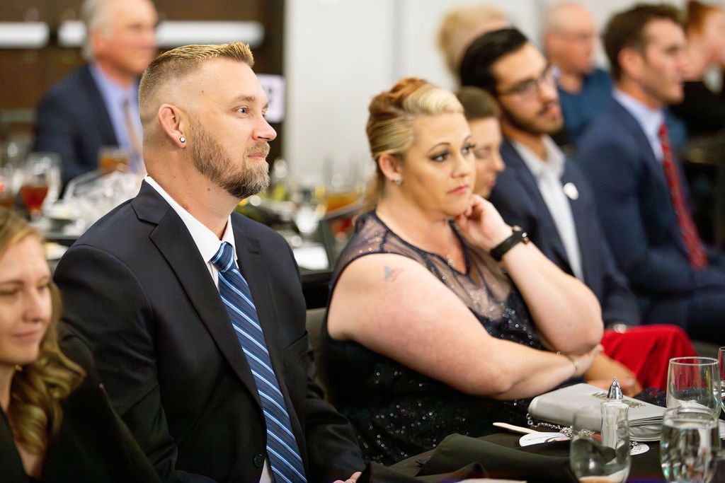 A group of formally dressed people sitting at tables during an indoor event, focused toward an off-screen speaker.