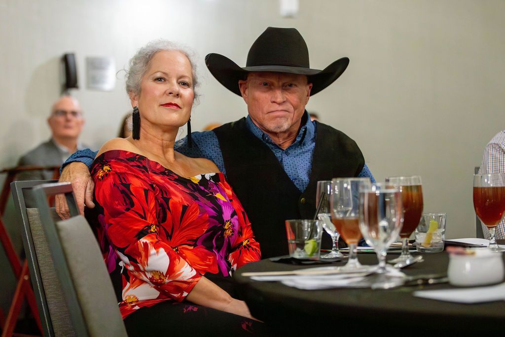 A man wearing a cowboy hat and a woman in a floral top sit at a table with drinks in a room with a background observer.