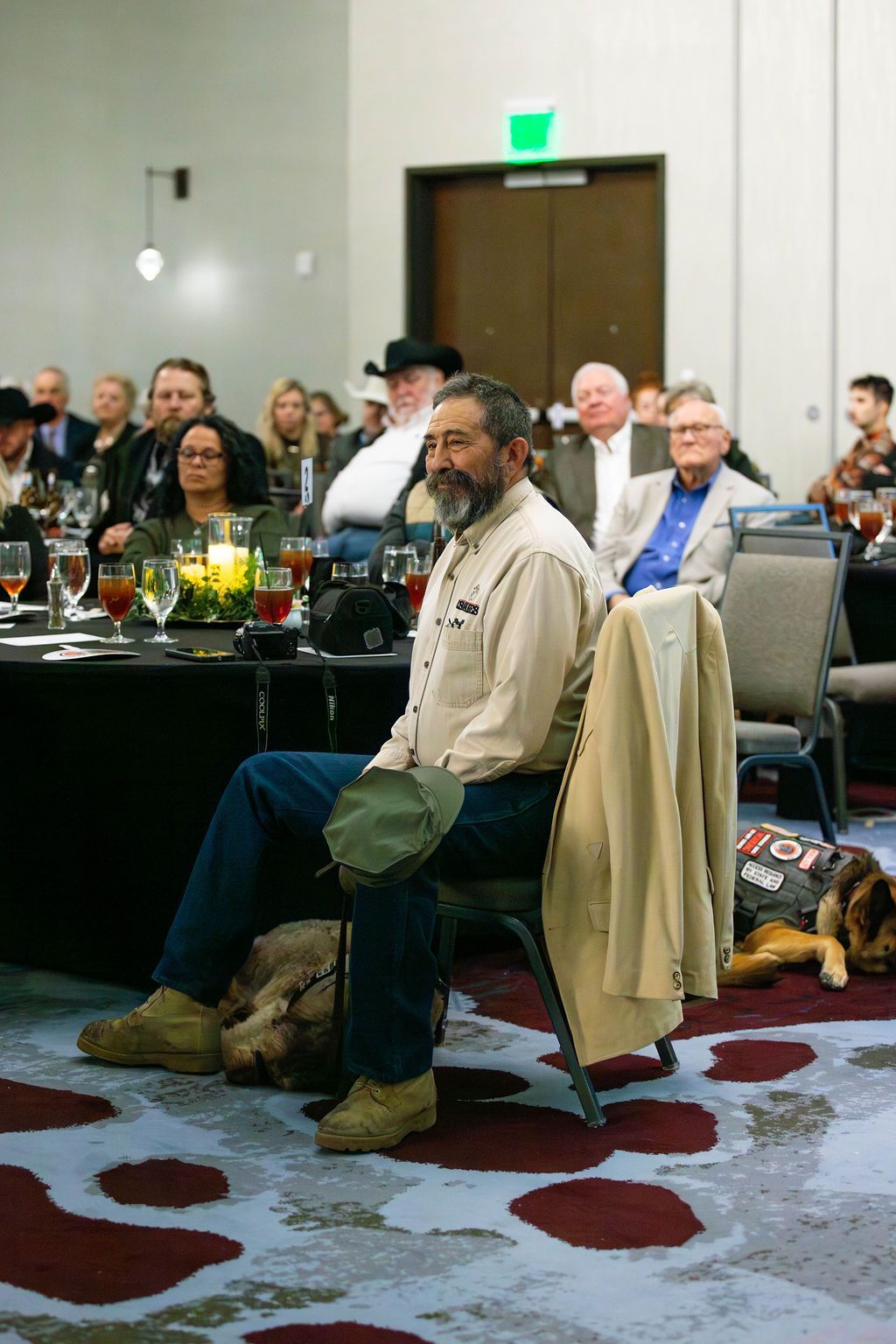 A bearded man in a tan shirt sits at a table in a conference room with a dog lying at his feet.