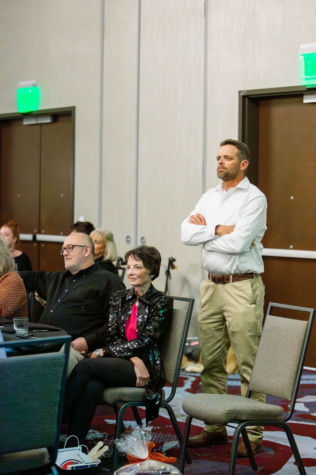 Attendees sit in chairs while a man stands with arms crossed in a conference room with green exit signs above doorways.