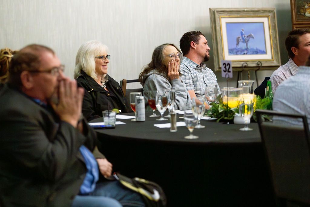 People sit at a black-clothed table in an indoor event space, with a framed painting on an easel in the background.