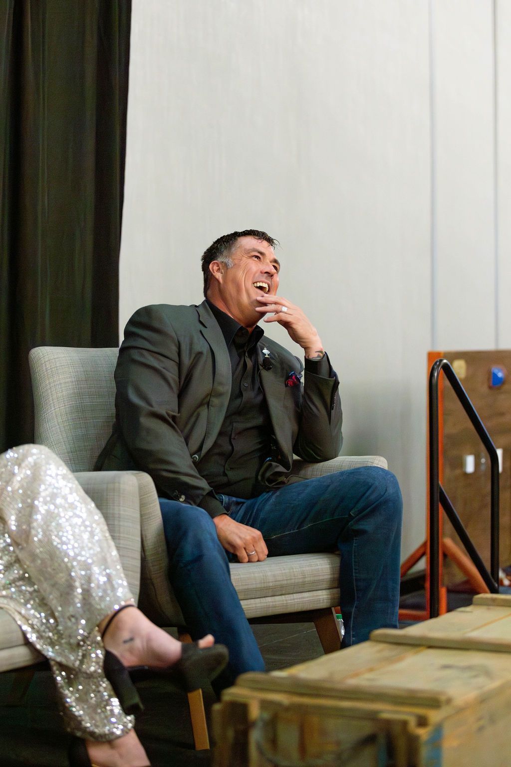 A smiling person in a dark blazer and blue jeans sits in a chair, hand to their chin, with a sequined pant leg in view.