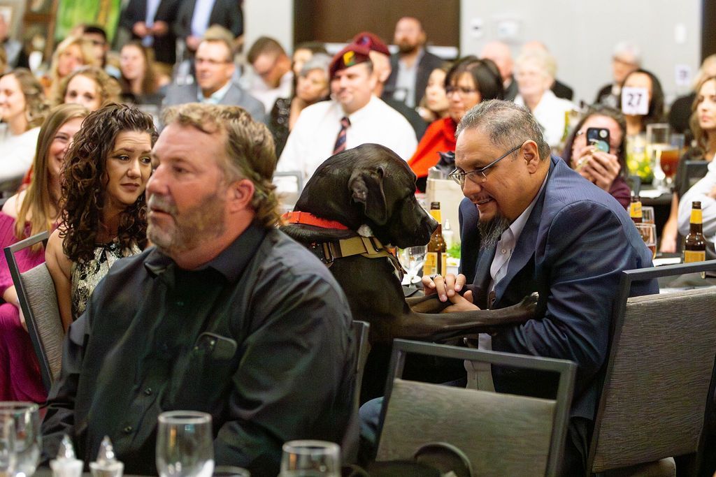 A black dog wearing a service harness sits between two men at a banquet table while people watch in a crowded room.