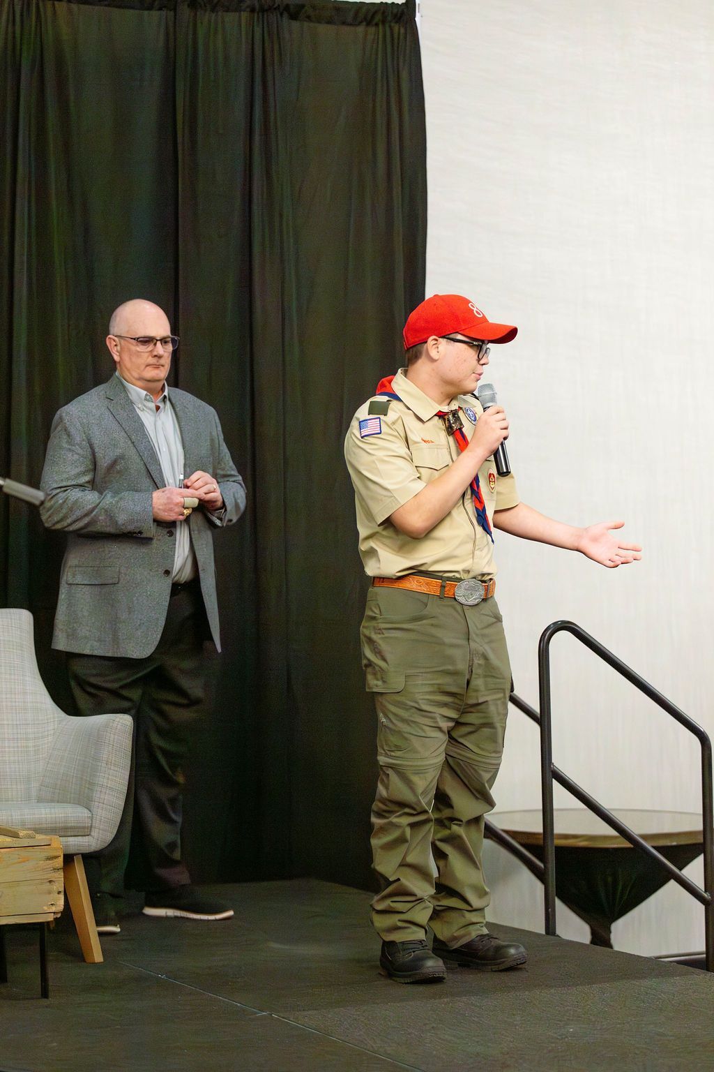 A scout in uniform speaks into a microphone on stage next to an adult in a gray suit and glasses.