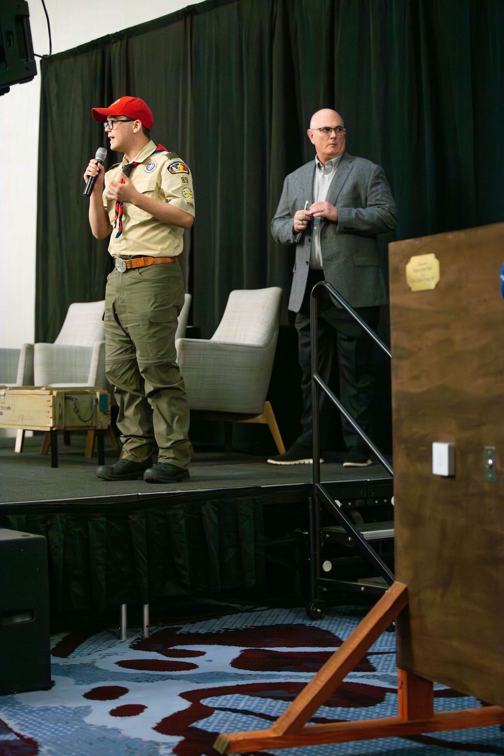A scout in a tan uniform and red cap speaks into a microphone on stage next to a man in a gray suit.