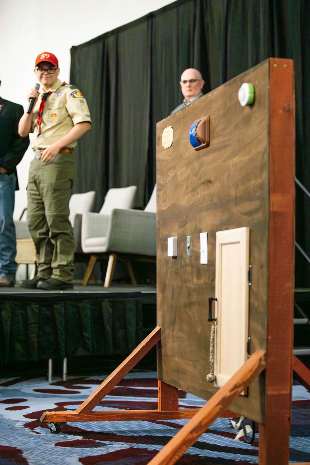 A scout speaks into a microphone next to a wooden training board with mounted hardware like a cabinet door and switches.