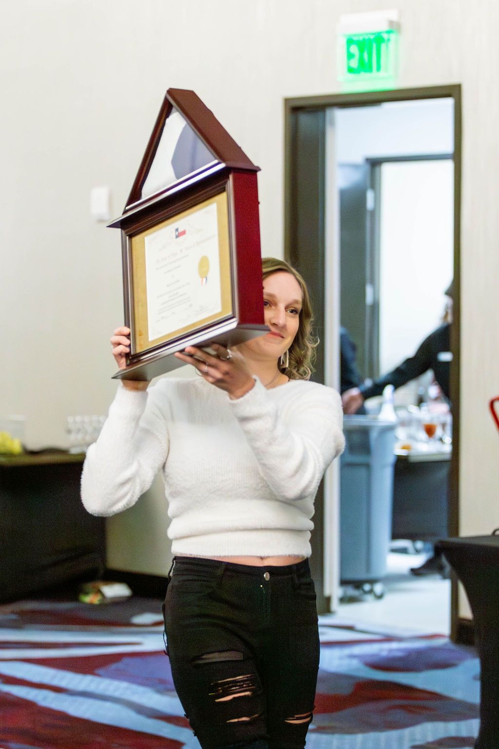 A person holding a framed, house-shaped award in front of their face, standing in an event room near an exit sign.