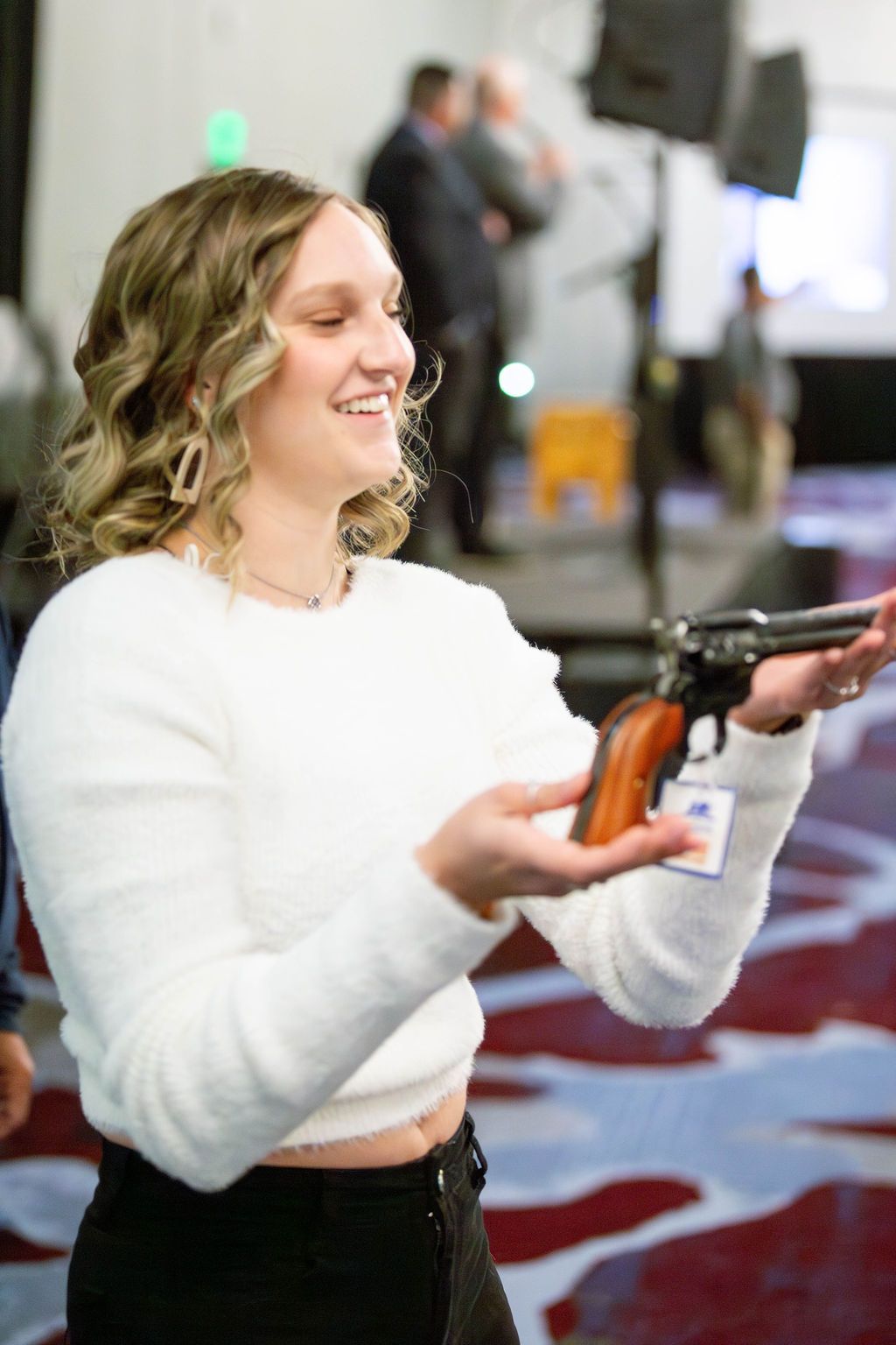 A person in a white sweater smiling while holding a vintage-style revolver with a wooden handle in a carpeted room.
