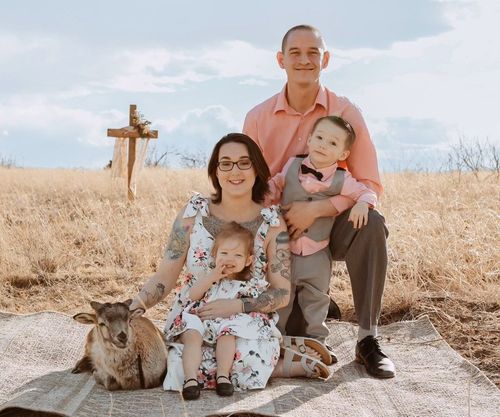 A family poses with a goat on a blanket in a field with a wooden cross in the background.