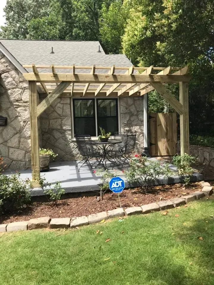 Wooden pergola over a patio with a table and chairs, near a stone house and garden.