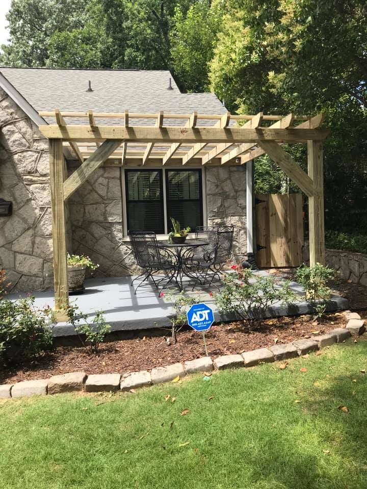 Pergola over a patio with table, chairs, and garden; stone house background.