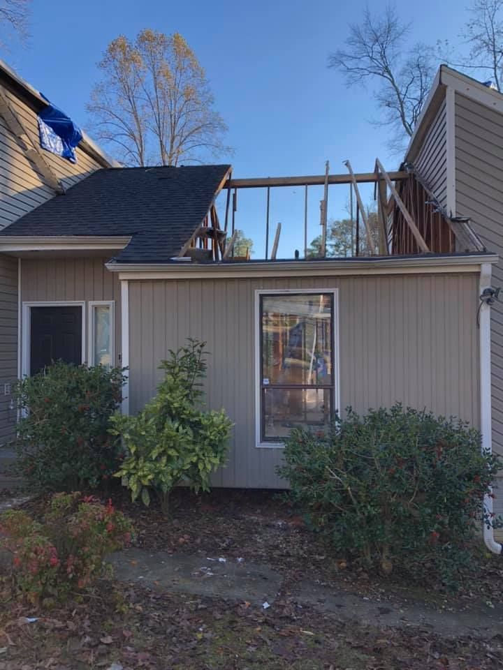 Exterior of a building with roof damage. Exposed wooden beams, siding, and overgrown bushes.
