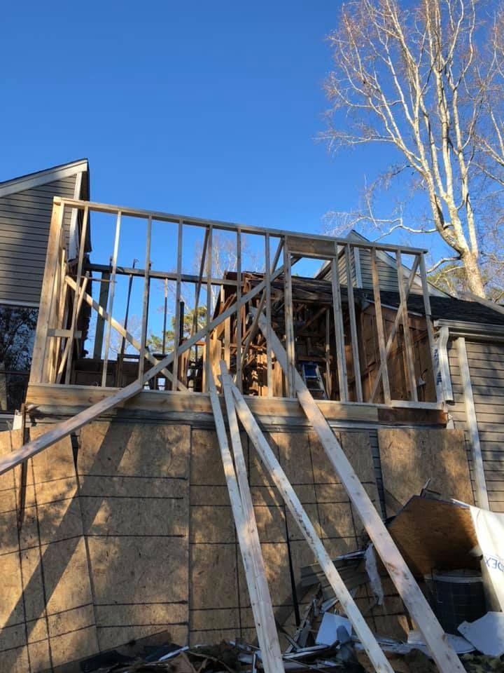 Exterior view of a house under construction with wood framing. Sunlight casts long shadows on the structure and ground.