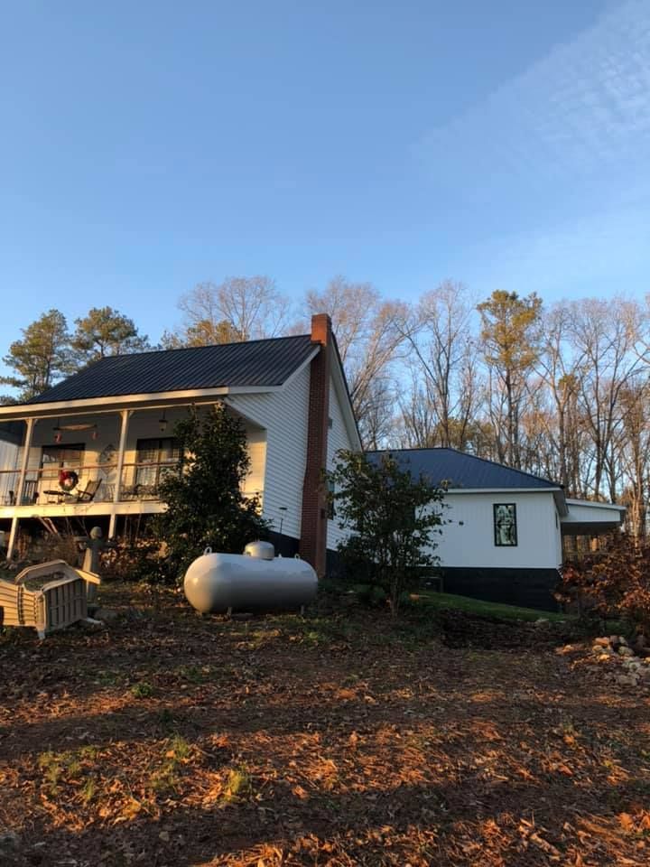 White farmhouse with a porch and chimney, large propane tank, trees, and a blue sky.