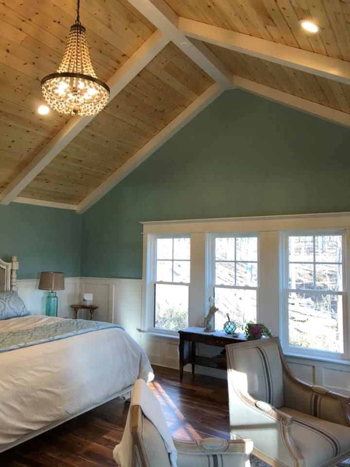 Bedroom with wood ceiling, teal walls, and chandelier; bed, windows, and chairs.