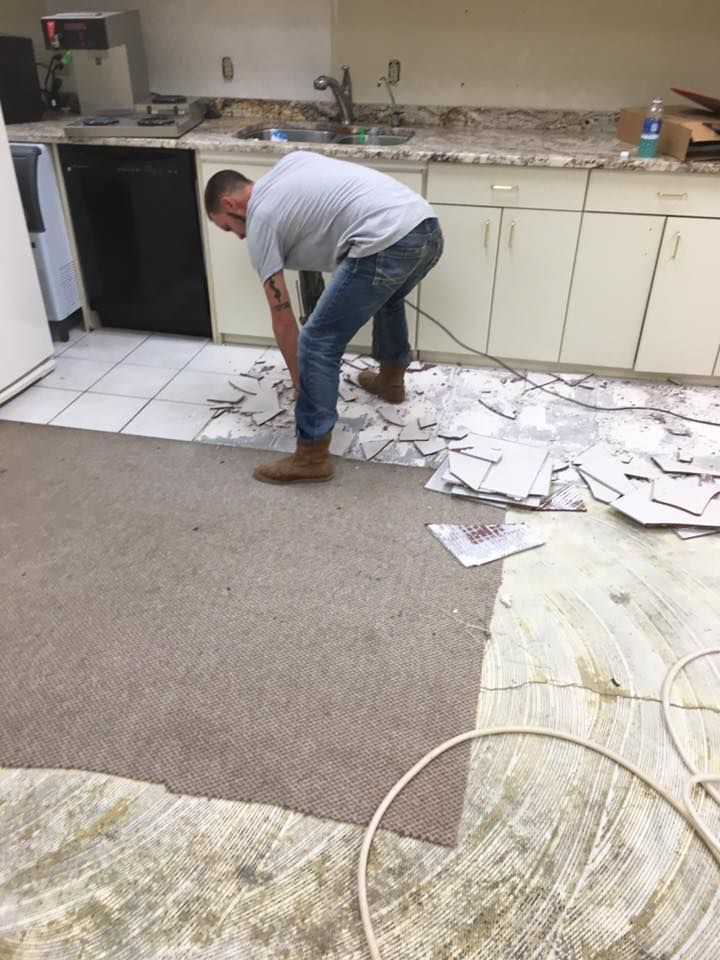 Man removing tile flooring in a kitchen. Torn tiles, tan carpet, and white cabinets are visible.