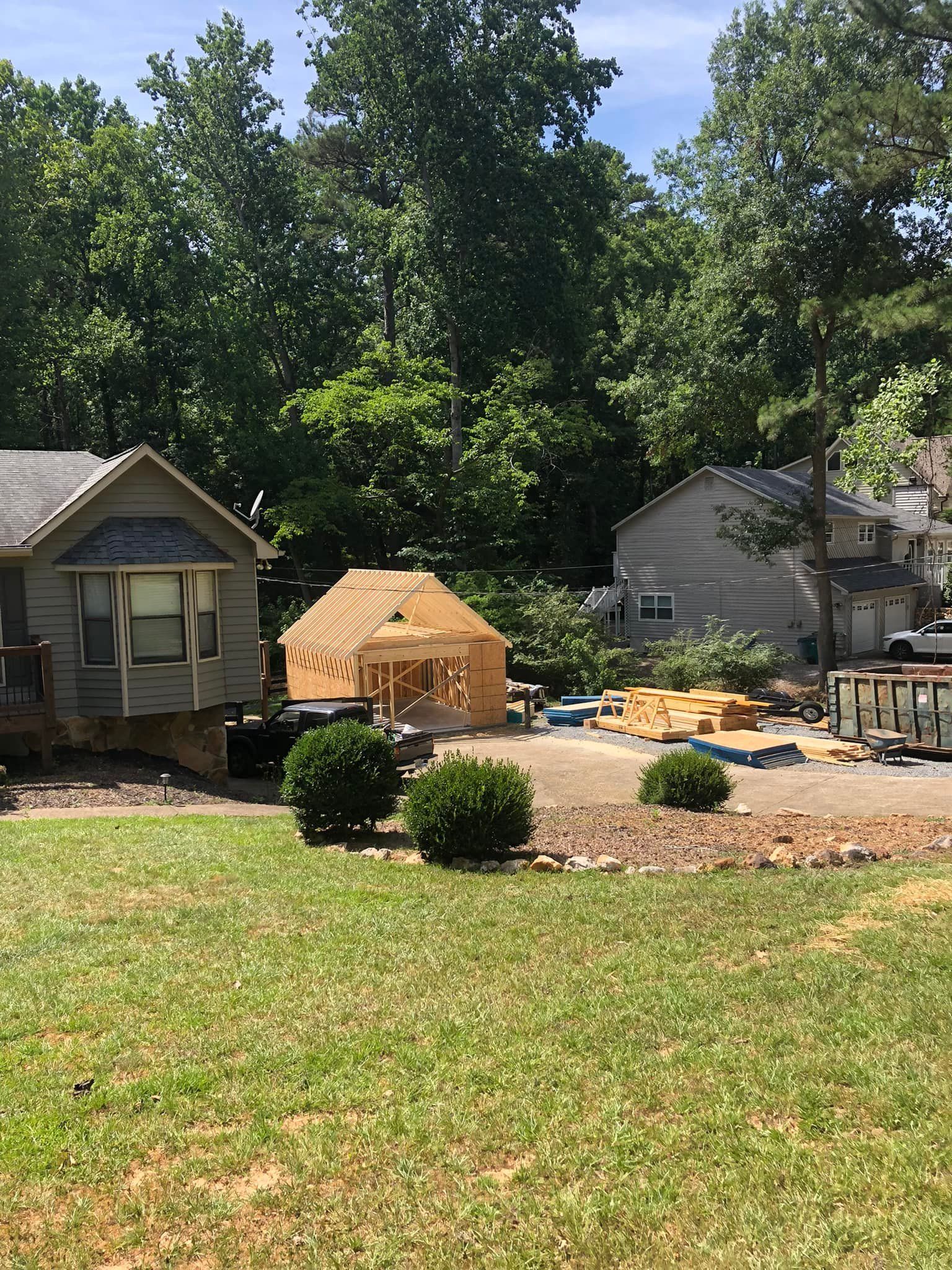 A wooden structure under construction between two houses, in a yard with green grass and trees in the background.