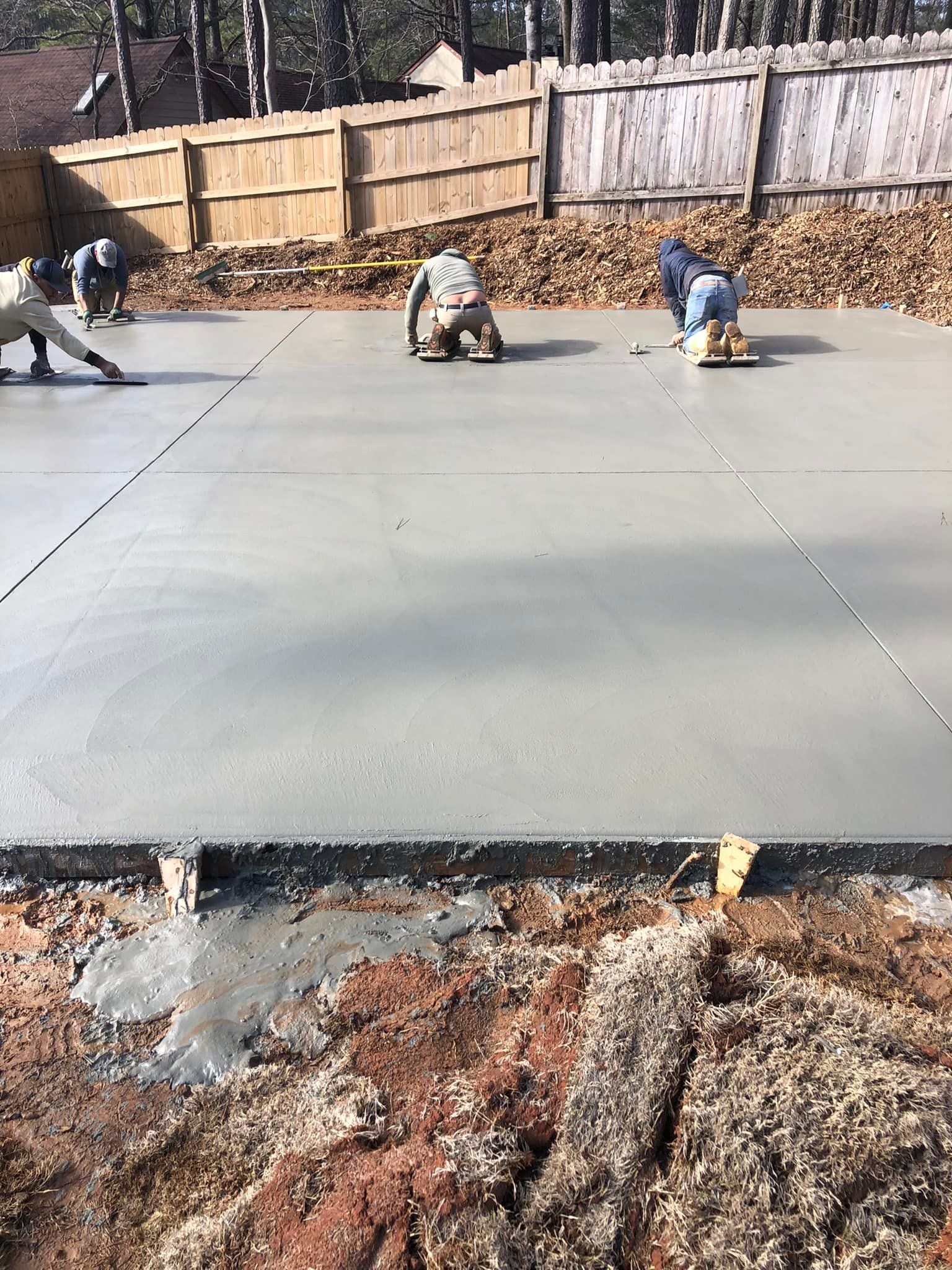 Workers smoothing fresh concrete slab; outdoor setting with wooden fence in background.