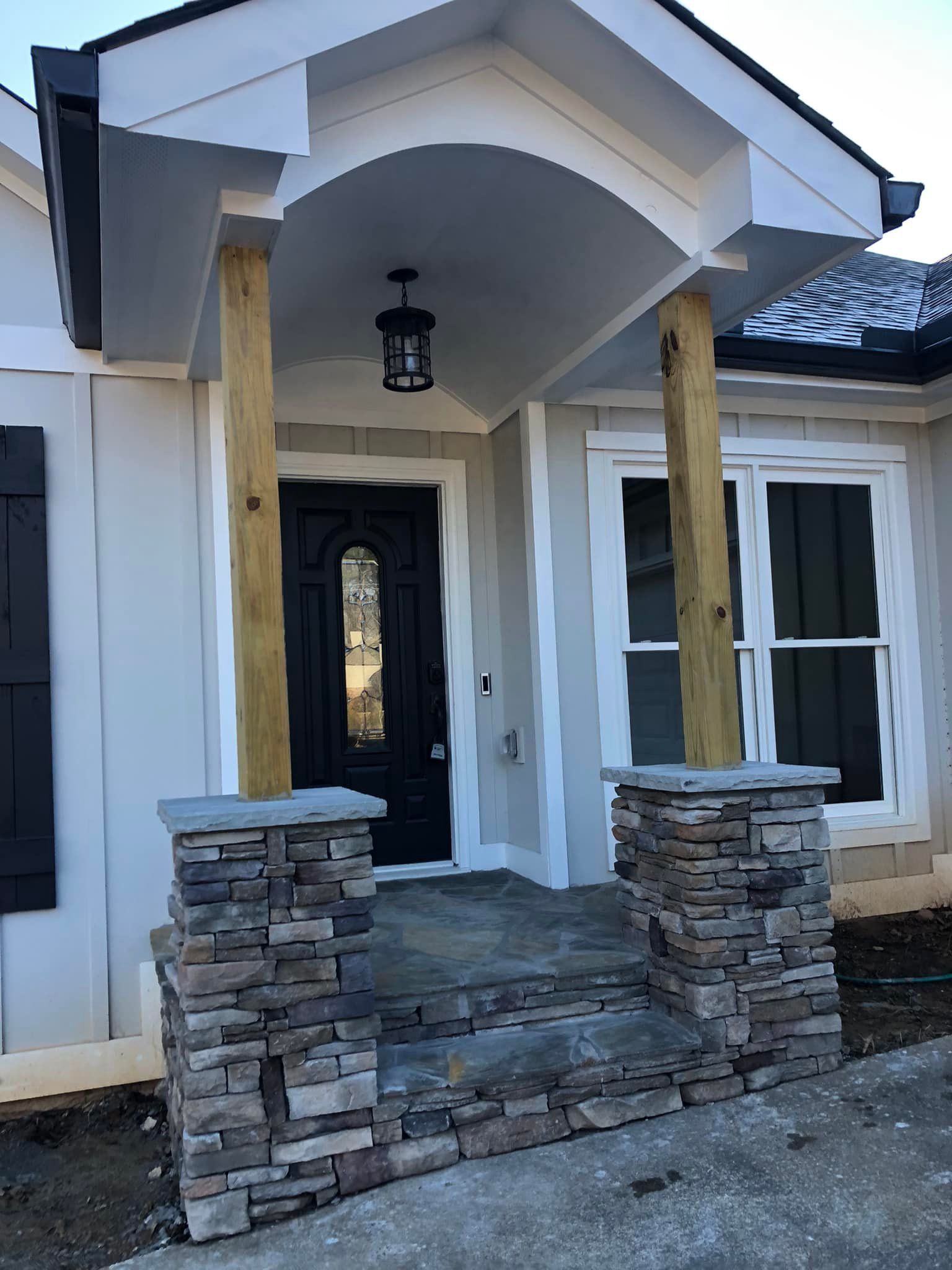 Front porch with stacked stone pillars, wooden posts, black door, and light fixture.