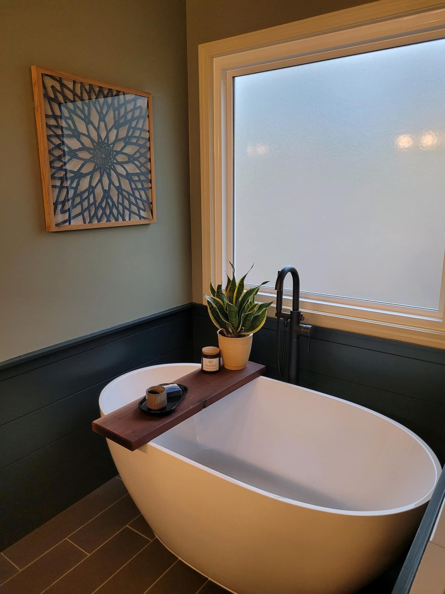 Freestanding white bathtub with wood tray, plant, and faucet in front of a frosted window.
