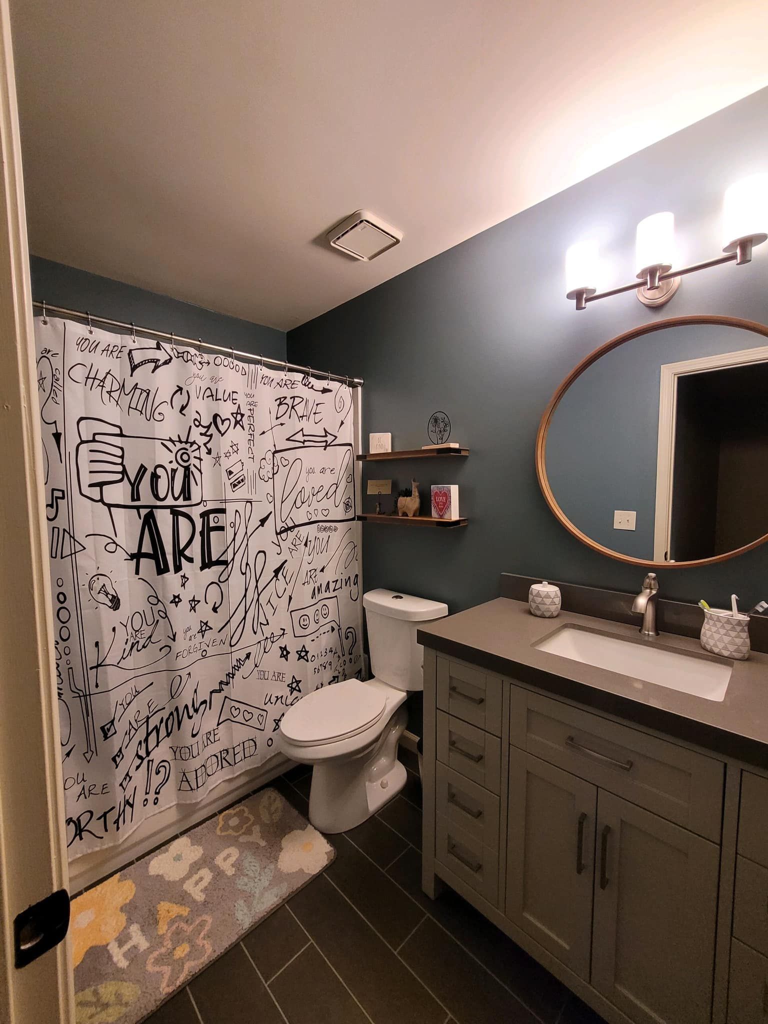 Bathroom with gray vanity, round mirror, and patterned shower curtain. Dark blue walls and dark tile floor.