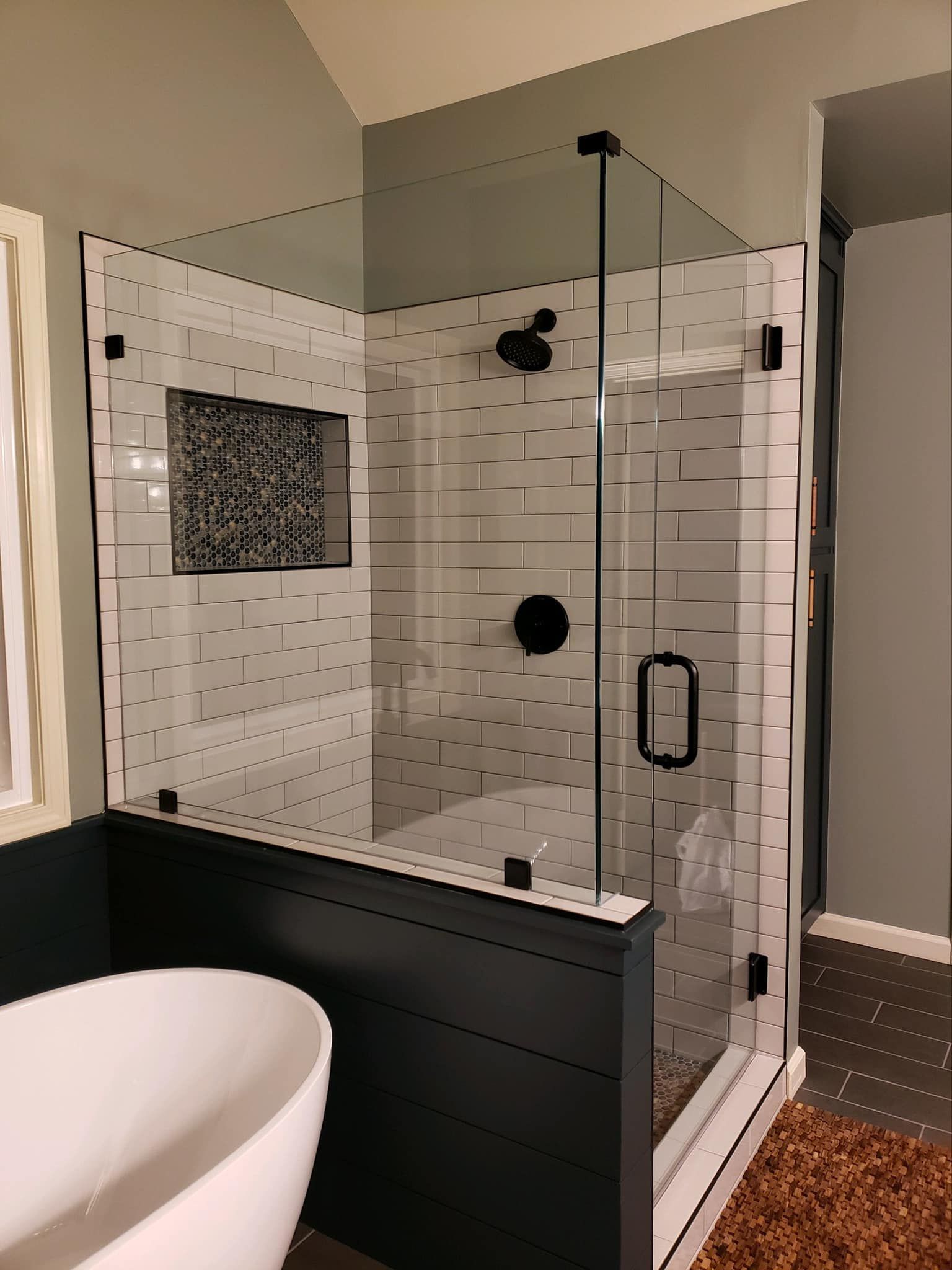 Modern bathroom with white tiled shower, black fixtures, and a glass door. A white tub sits in front.