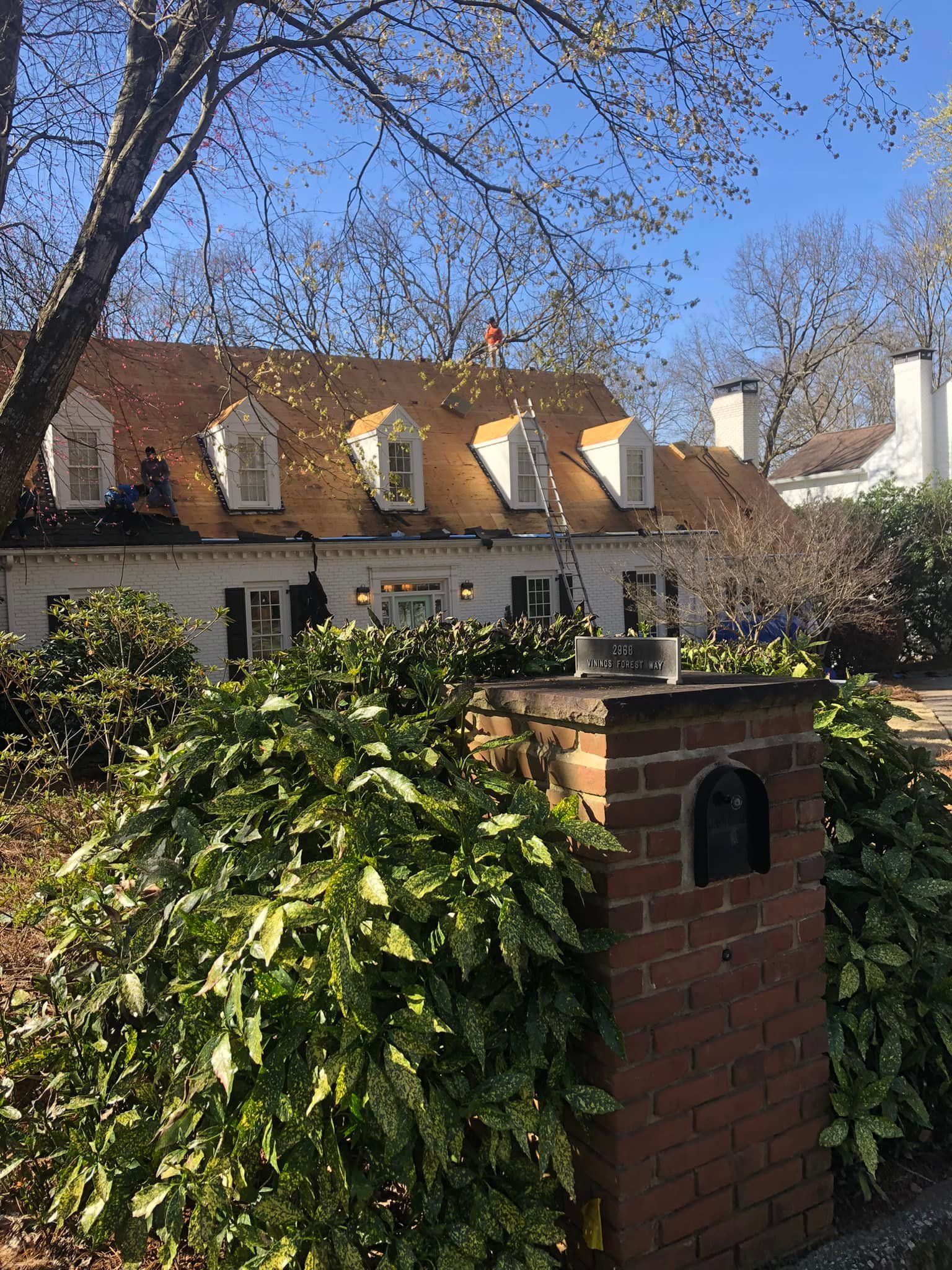 White house with dormer windows and brick column, surrounded by greenery under a blue sky.
